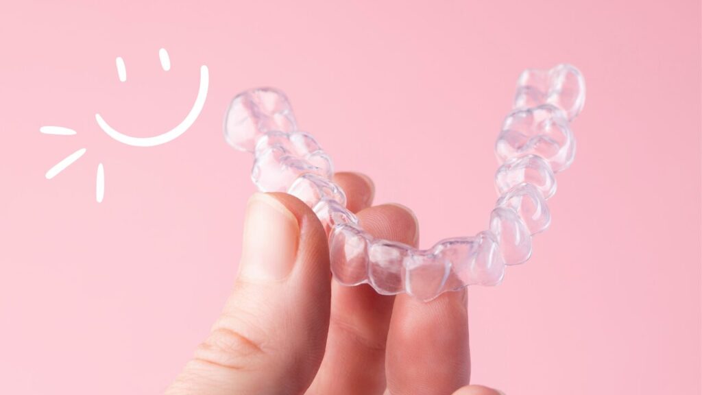 ピンクの背景。透明な矯正用マウスピースを持っている手もとの画像。